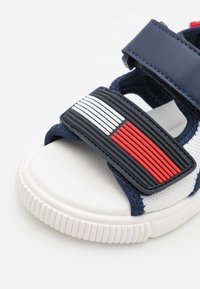 Marinblå sandal med en texturerad gummisnodd med röda och vita detaljer, en vit texturerad fotbädd och en räfflad vit sula.