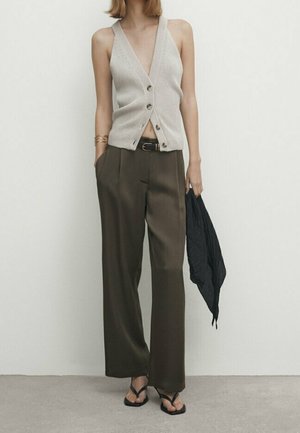 Femme portant un top sans manches en maille beige boutonné, un pantalon large olive, des tongs noires, tenant une écharpe noire dans une main.