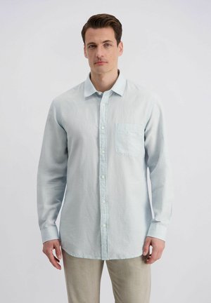 Man met bruin haar die een lichtblauw overhemd met knopen en beige broek draagt, staand tegen een effen witte achtergrond, vooruit gericht.