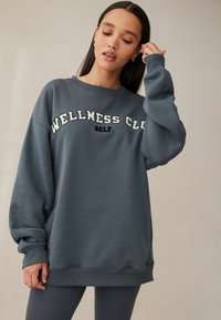 Šedá oversized mikina s nápisom "WELLNESS CLUB" a "SELF." v kontrastnej bielej farbe; rebrované manžety a spodný lem. Hladká textúra látky.