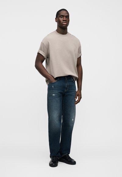 Μπεζ μπλουζάκι με ρεβερ μανίκια, μπλε φθαρμένο τζιν με ίσια γραμμή και μαύρα αθλητικά παπούτσια. Το σύνολο έχει χαλαρή εφαρμογή και ελαφρύ ξεθώριασμα στο τζιν.