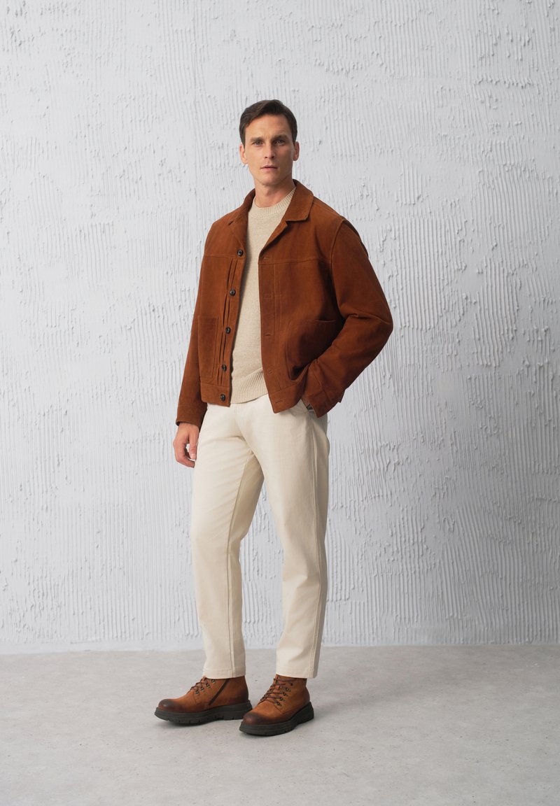 Brązowa zamszowa kurtka z guzikami, beżowy sweter, kremowe spodnie i brązowe buty trekkingowe. Tekstura szara w tle. Stylizacja casualowa.