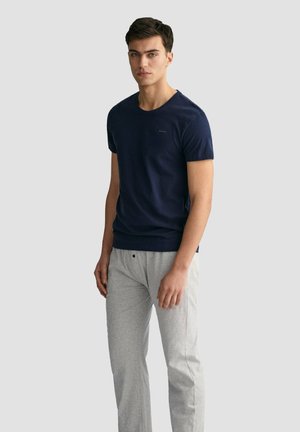 Młody mężczyzna stoi, ubrany w granatowy t-shirt z krótkim rękawem i jasnoszare spodnie na jednolitym jasnoszarym tle.