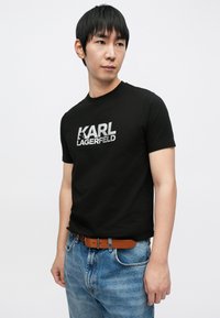 Melna īsroku T-krekls ar sudraba logotipa apdruku. Modelis valkā to kopā ar zilām džinsām un brūnu siksnu, stāvot neitrālā pozā.