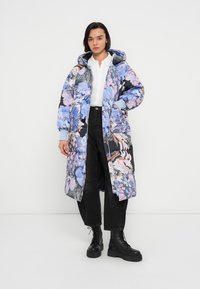 Manteau matelassé avec un motif floral en bleu et rose, doté d'une capuche, de poignets côtelés et d'une fermeture éclair. Porté avec un pantalon noir et des bottes.
