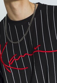 Czarny t-shirt z krótkim rękawem z pionowymi białymi paskami, z czerwonym wyszywanym logo. Na linii szyi widoczny srebrny naszyjnik z łańcuszka.