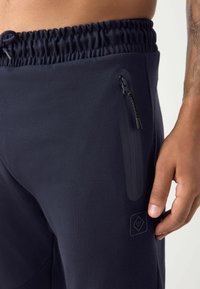 Los pantalones atléticos en color navy cuentan con una cintura elástica, un bolsillo lateral con cremallera y una discreta solapa, además de un detalle con el logo cerca del bolsillo.