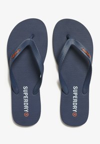 Blå flip-flops med teksturerede såler, der har et fiskebensmønster og et kontrastfarvet orange logo på stropperne og foden.