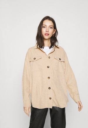 Veste chemise en velours côtelé beige avec deux poches avant et de grands boutons marron, arborant une coupe décontractée et un col de chemise, associée à un pantalon noir.