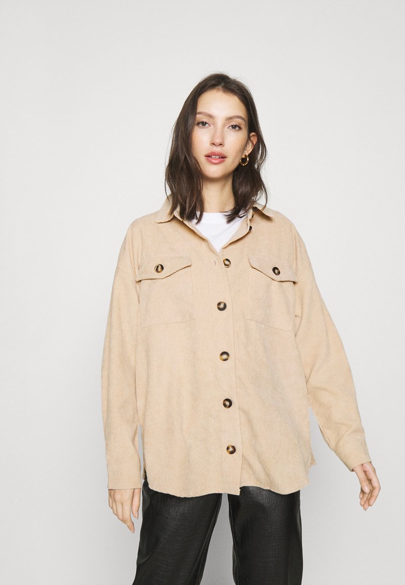 Beige Kordjacke mit zwei vorderen Taschen und großen braunen Knöpfen, im lässigen Schnitt und mit Hemdkragen, kombiniert mit schwarzen Hosen.