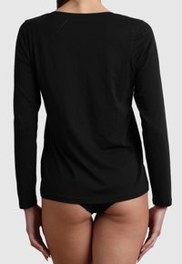 Donna che indossa un top nero a maniche lunghe e biancheria intima nera, voltata di spalle con le mani rilassate ai lati, su uno sfondo neutro.