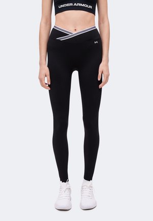 Γυναίκα φοράει μαύρα κολάν Under Armour με σταυρωτή μέση, μαύρο αθλητικό σουτιέν και λευκά αθλητικά παπούτσια, στέκεται όρθια.