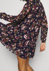 Robe fleurie bleu marine à manches longues. Tissu léger orné de fleurs roses, rouges et vertes. Silhouette évasée avec des détails froncés.