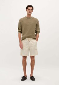 Shorts cargo beiges avec poches latérales, associés à un pull vert olive côtelé. Le modèle se tient sur un fond neutre, portant des chaussures sombres.