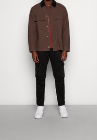 Man wearing brown jacket over red shirt, black cargo pants, and white sneakers standing against a plain background.