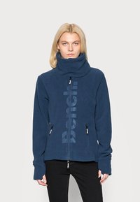Navy fleece jacket with high collar, full zip, and two zippered pockets. Features large logo print on front and thumbhole cuffs.