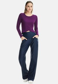 Purple long-sleeve top with a twist at the front, paired with navy wide-leg pants featuring side pockets and a smooth texture. White shoes.