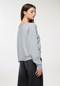 Maglione grigio chiaro in tessuto morbido, con scollo ampio, vestibilità rilassata e dettagli con bottoni sulla spalla. Abbinato a jeans scuri.
