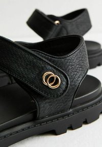Svarta sandaler med texturerade syntetiska remmar och en kraftig sula. Guldiga cirkulära detaljer ger extra finess åt remdesignen.