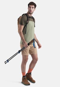 Uomo che indossa una camicia verde chiaro, pantaloni corti beige, calze arancioni e scarponi da trekking marroni, mentre tiene in mano dei bastoni da trekking neri e blu, con uno zaino beige.