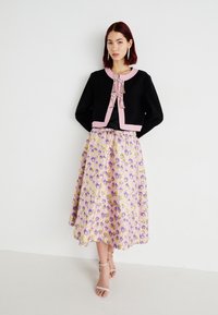 Jachetă scurtă neagră cu trimuri roz și ciucuri decorative, asortată cu o fustă cu model floral pastel. Sandale simple completează look-ul.