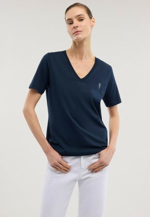 Kobieta ubrana w granatowy t-shirt z krótkim rękawem i dekoltem w serek oraz białe spodnie, stoi z rękami blisko kieszeni na jednolitym tle.