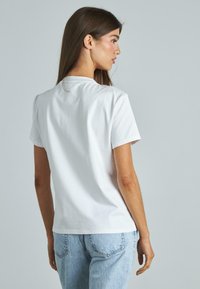 Camiseta blanca de algodón con mangas cortas, cuello redondo y textura suave. Presenta un ajuste regular, mostrada desde la parte de atrás con jeans de mezclilla azules.