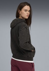 Dunkelgrauer, übergroßer Kapuzenpullover aus weichem, strukturiertem Stoff mit gefütterter Kapuze. Verfügt über eine Kängurutasche und gerippte Bündchen, getragen über einem weißen Shirt.