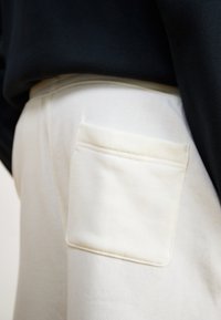 Vita byxor med en texturerad yta har en tydlig, fyrkantig patchficka. Midjebandet är delvis täckt av ett svart plagg ovanför.