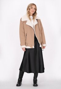 Beige Kunstlederjacke mit einem weißen Kunstfellkragen, seitlichen Reißverschlüssen und Schnallen-Details, kombiniert mit einem schwarzen Satinklied und schwarzen Stiefeln.