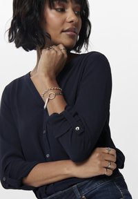 Blusa blu navy con scollo a V e dettagli con bottoni. Presenta maniche arrotolate con chiusure. Gli accessori includono bracciali d'argento a strati e anelli.