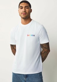 Hvid bomulds t-shirt med korte ærmer, med et multikolor "be you" tekstlogo på brystet. Sammen med blå denim jeans.