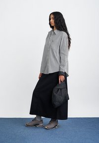 Schwarz-weiß gestreiftes übergroßes Hemd, langer schwarzer Rock, graue Schuhe, hält eine gesteppten schwarzen Tasche, vor einem schlichten weißen Hintergrund.