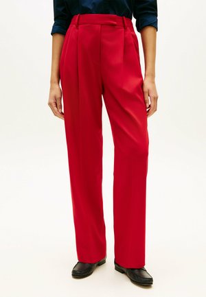 Person iført højttaljede, røde bukser med vide ben og folder, kombineret med en mørkeblå skjorte og sorte loafers, stående på en hvid baggrund.