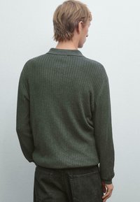Person med kort blont hår vendt væk, iført en ribbet mørkegrøn langærmet skjorte og sorte jeans mod en ensfarvet lys væg.