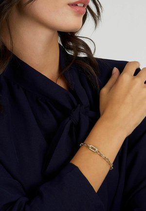 Bracciale in oro con maglie texture e un centro ovale, indossato su un polso con pelle abbronzata, abbinato a una blusa scura annodata.