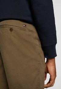 Beige Chinos mit glatter Textur, ausgestattet mit einer hinteren Tasche mit Knopf und einem kleinen, in Rot, Weiß und Blau gestickten Logo.