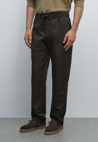 Pantalons amples marron avec une taille élastique et cordon de serrage, confectionnés en tissu léger, assortis à des chaussures en suede foncé.