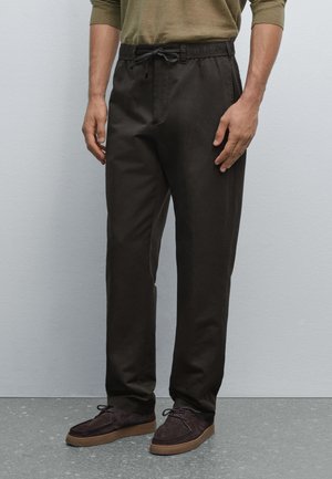 Bruine, loszittende broek met een elastische tailleband en trekkoord, gemaakt van lichtgewicht stof, gecombineerd met donker suede schoenen.