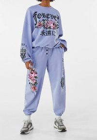 Survêtement violet clair avec des imprimés floraux. Le haut à manches longues comporte un texte, tandis que le pantalon a un cordon de serrage et des poignets. Le matériau semble doux.