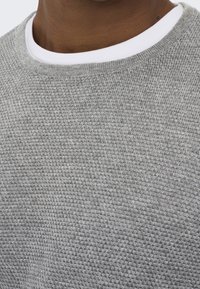 Maglione grigio lavorato a maglia con un motivo testurizzato e colletto rotondo bianco. Vista ravvicinata che mostra i dettagli del tessuto e delle cuciture.