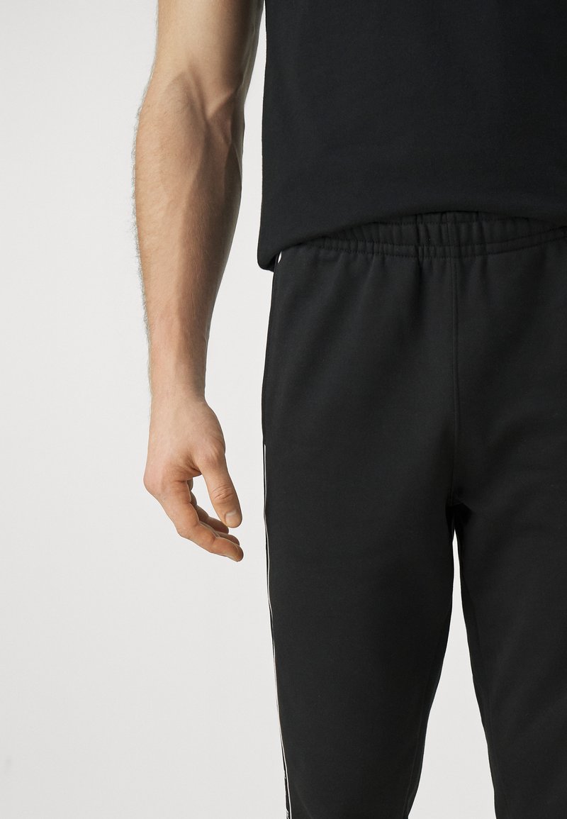 Czarne dresy z elastycznym pasem, z białymi paskami po bokach. Zbliżenie męskiej ręki umieszczonej obok pasa.