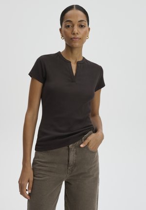 Vrouw met donker haar in een lage knot draagt gouden creolen, een zwart top met korte mouwen en een kleine V-hals, en grijze broek met één hand in de zak.