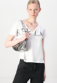 White V-neck T-shirt with "REPL" printed in gray. Worn with black jeans featuring metallic dot accents and holding a silver metallic shoulder bag.
