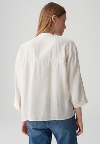 Camicia ampia di colore crema, realizzata in tessuto leggero, con maniche a tre quarti e aperture laterali; texture liscia, design minimale.