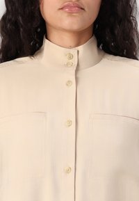 Beige collared shirt featuring a button-down front, two chest pockets, smooth texture, and subtle diagonal pattern in the fabric.