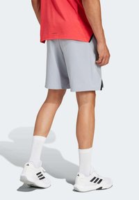 Pantalones cortos deportivos gris claro con aberturas laterales, combinados con zapatillas deportivas blancas con rayas negras y calcetines blancos hasta el tobillo. Camiseta roja visible.