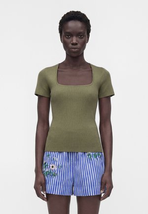 Γυναίκα φορώντας ελαφρώς ριγωτό τοπ ελιάς με κοντά μανίκια και μπλε-άσπρο ριγωτό σορτς με κεντήματα λουλουδιών, στέκεται μπροστά σε απλό φόντο.