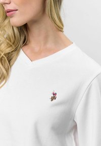 Biely sveter s V-výstrihom, vyrobený z mäkkej tkaniny. Obsahuje vyšívané logo na hrudi v živých farbách.