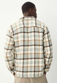Homme portant une veste à carreaux beige, marron et bleu clair, se tenant les mains dans les poches, tourné vers le fond uni.